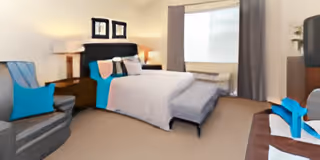 A cozy bedroom featuring a neatly made bed with white and blue bedding, a wooden nightstand with a lamp, a comfortable armchair with a blue cushion, a window with gray curtains, and framed artwork on the wall.