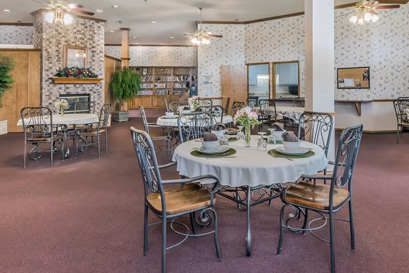 A dining area in a senior living facility with round tables covered in white tablecloths, set with plates, bowls, glasses, and napkins. The room has carpeted floors, floral wallpaper, ceiling fans with lights, and a fireplace with a floral arrangement. There are bookshelves and a serving window in the background.