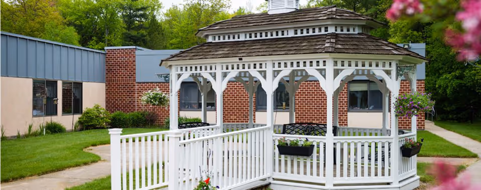 White wooden gazebo with benches inside, surrounded by green grass and a paved walkway, with a brick and beige building in the background and trees behind it.