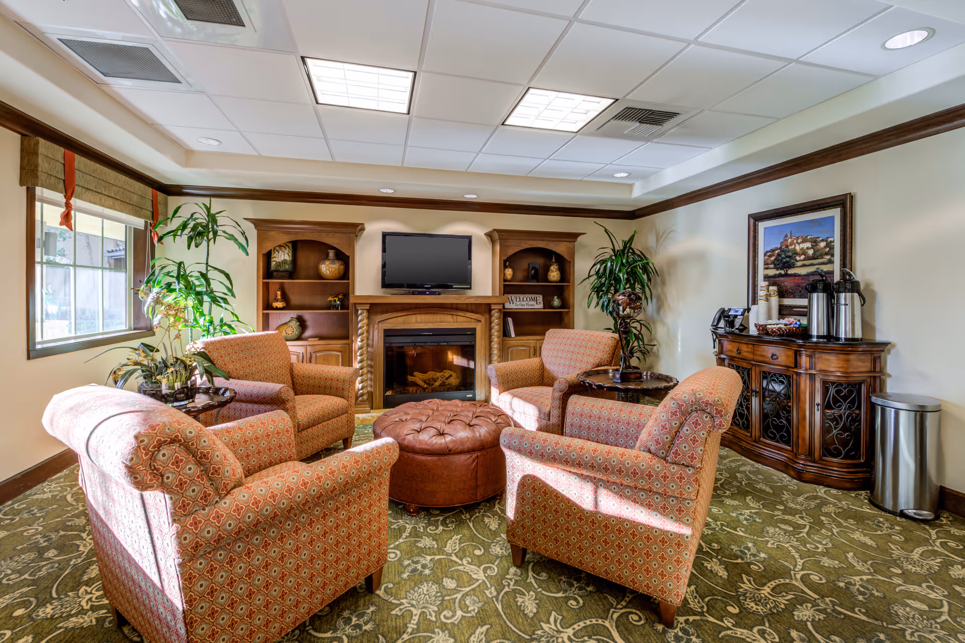 A cozy living room area with four patterned armchairs arranged around a round brown leather ottoman. There is a fireplace with a TV mounted above it, flanked by wooden shelves with decorative items. A large window with a brown valance lets in natural light. A wooden sideboard with coffee dispensers and cups is against the wall, next to a trash can. The room has a green patterned carpet and cream-colored walls with dark wood trim.