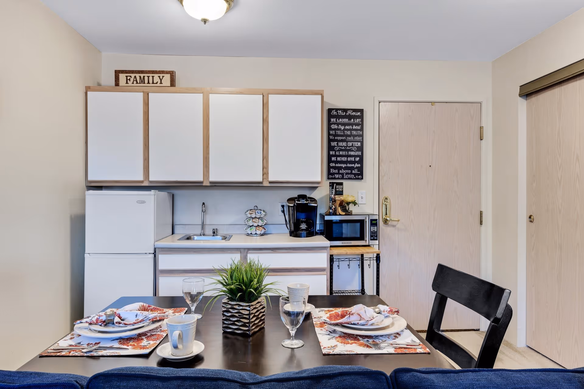 A small kitchen area with white cabinets, a mini refrigerator, a sink, a coffee maker, and a microwave on a wooden cart. In front of the kitchen is a dining table set for two with plates, glasses, cups, and a small potted plant centerpiece. There is a sign on top of the cabinets that says 'FAMILY' and a decorative sign with text on the wall next to a closed door.