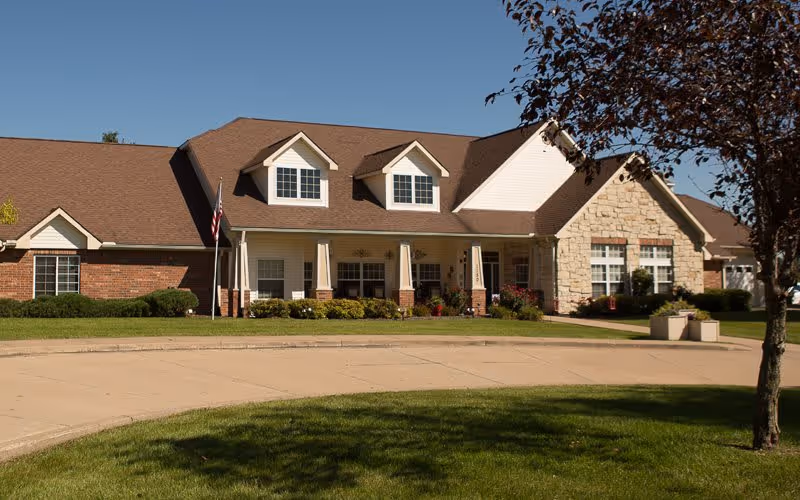 Front exterior view of a single-story senior living facility building with a brown roof, beige siding, brick and stone accents, a covered porch with columns, an American flag, and a curved driveway surrounded by green grass and trees.