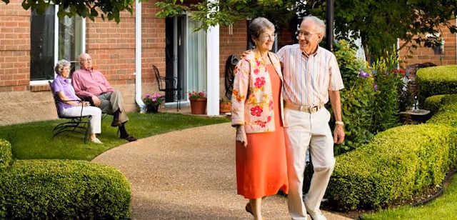 Two elderly women and one elderly man sitting on benches outside a brick building, while another elderly couple walks arm in arm along a paved garden path surrounded by green bushes and plants.