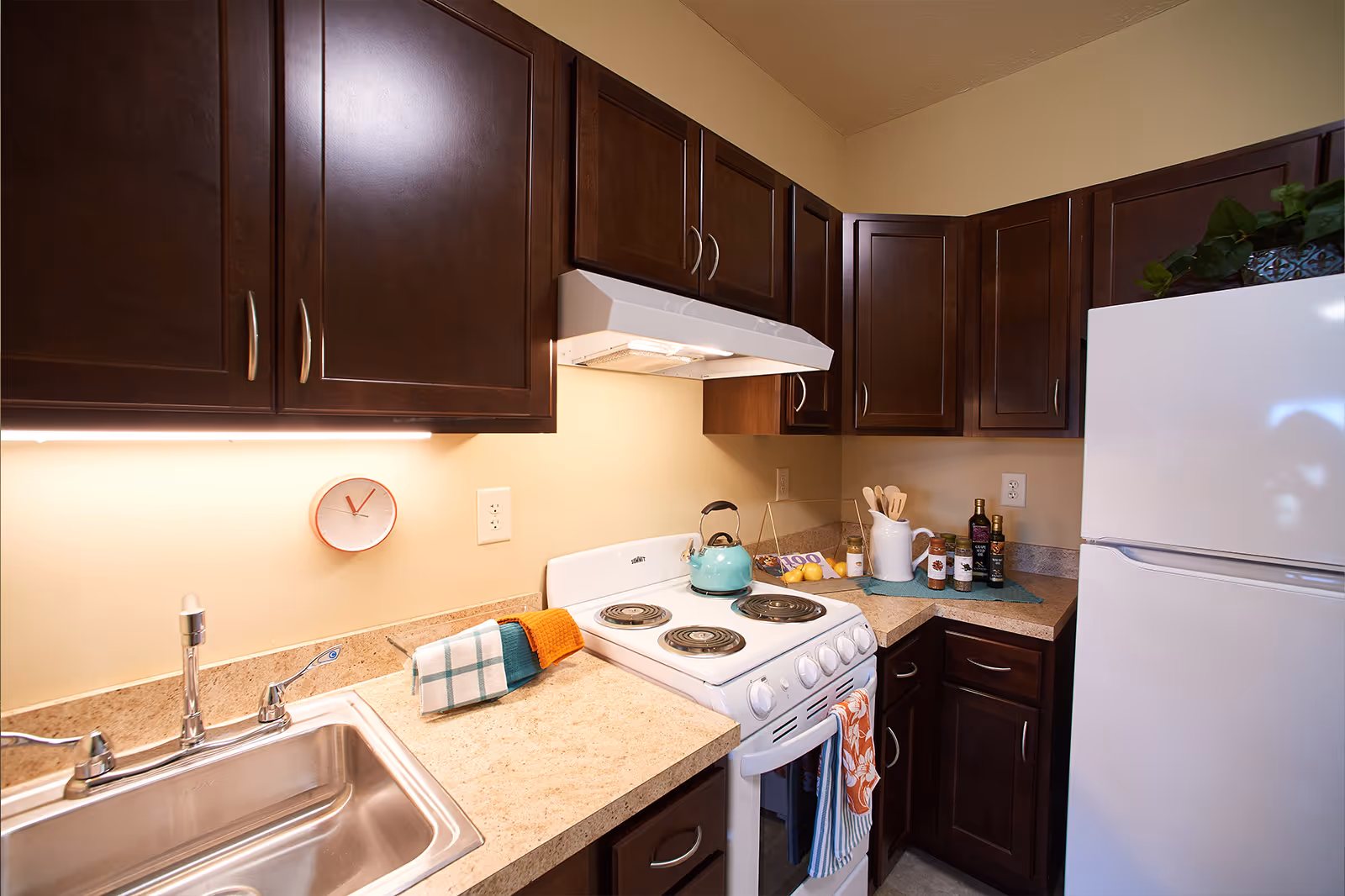 Compact kitchen with dark wood cabinets, a white stove and refrigerator, stainless sink, and countertop accessories.