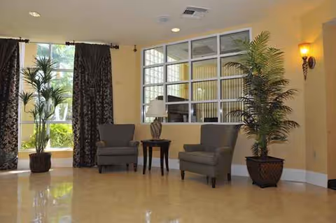 Spacious indoor lounge with two gray armchairs, a small table, lamps, and potted plants.