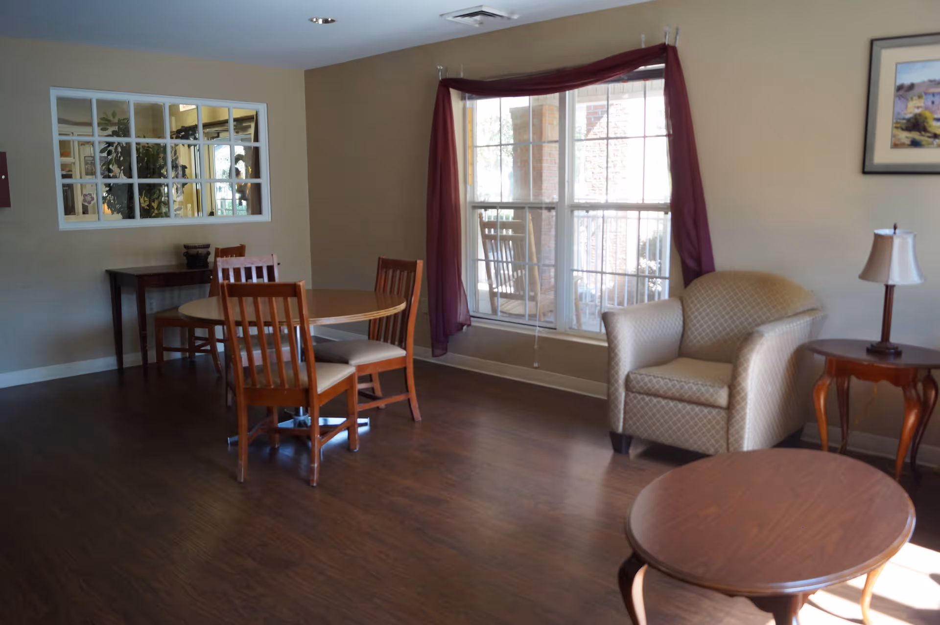 A bright communal living area with a round dining table and wooden chairs, an armchair by a window with purple curtains, and wooden side tables.