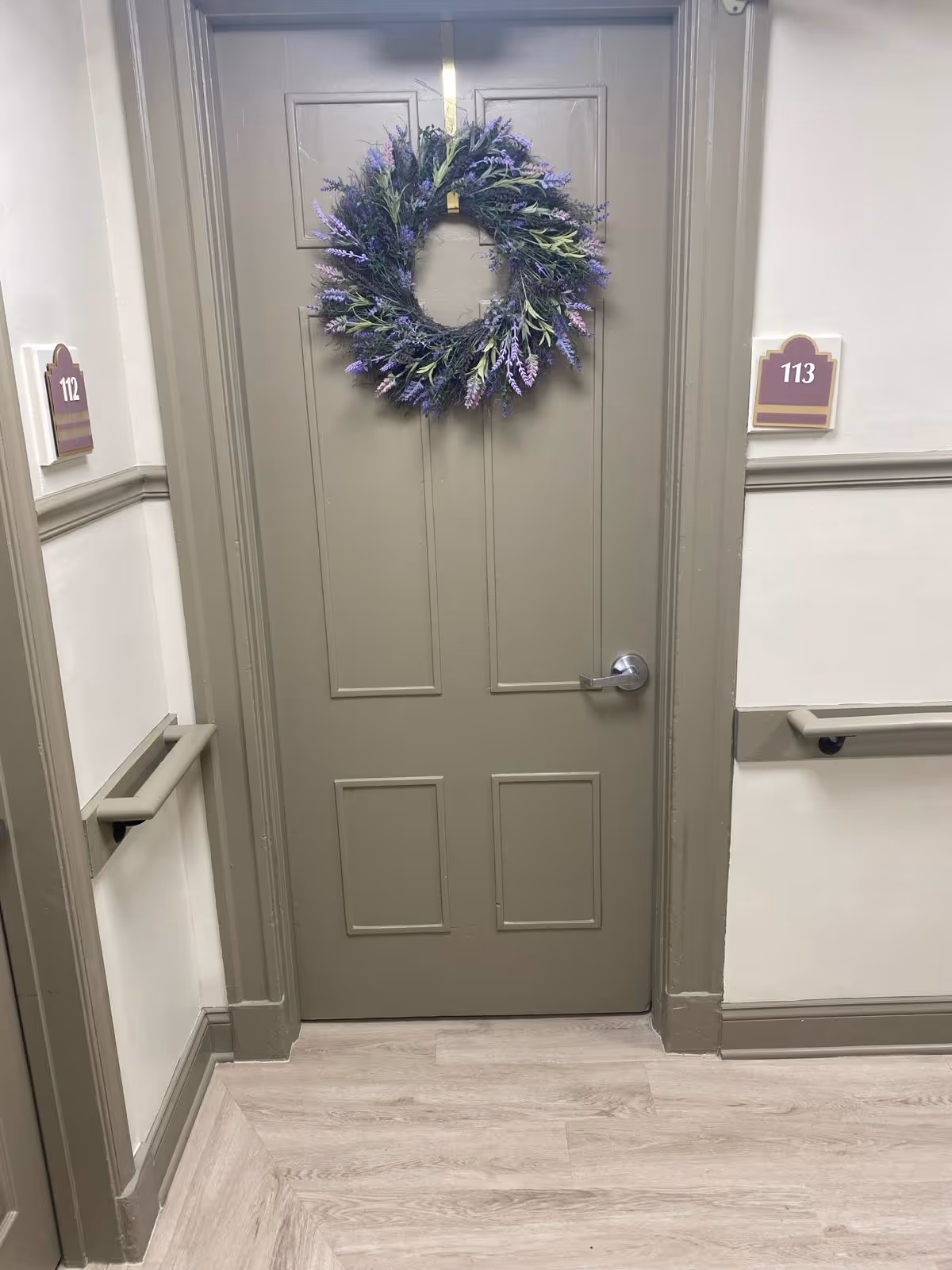 A closed beige door decorated with a wreath made of purple and green foliage. The door is located in a hallway with light-colored walls and wood flooring. Room number signs 112 and 113 are visible on the walls beside the door, along with handrails on both sides of the hallway.
