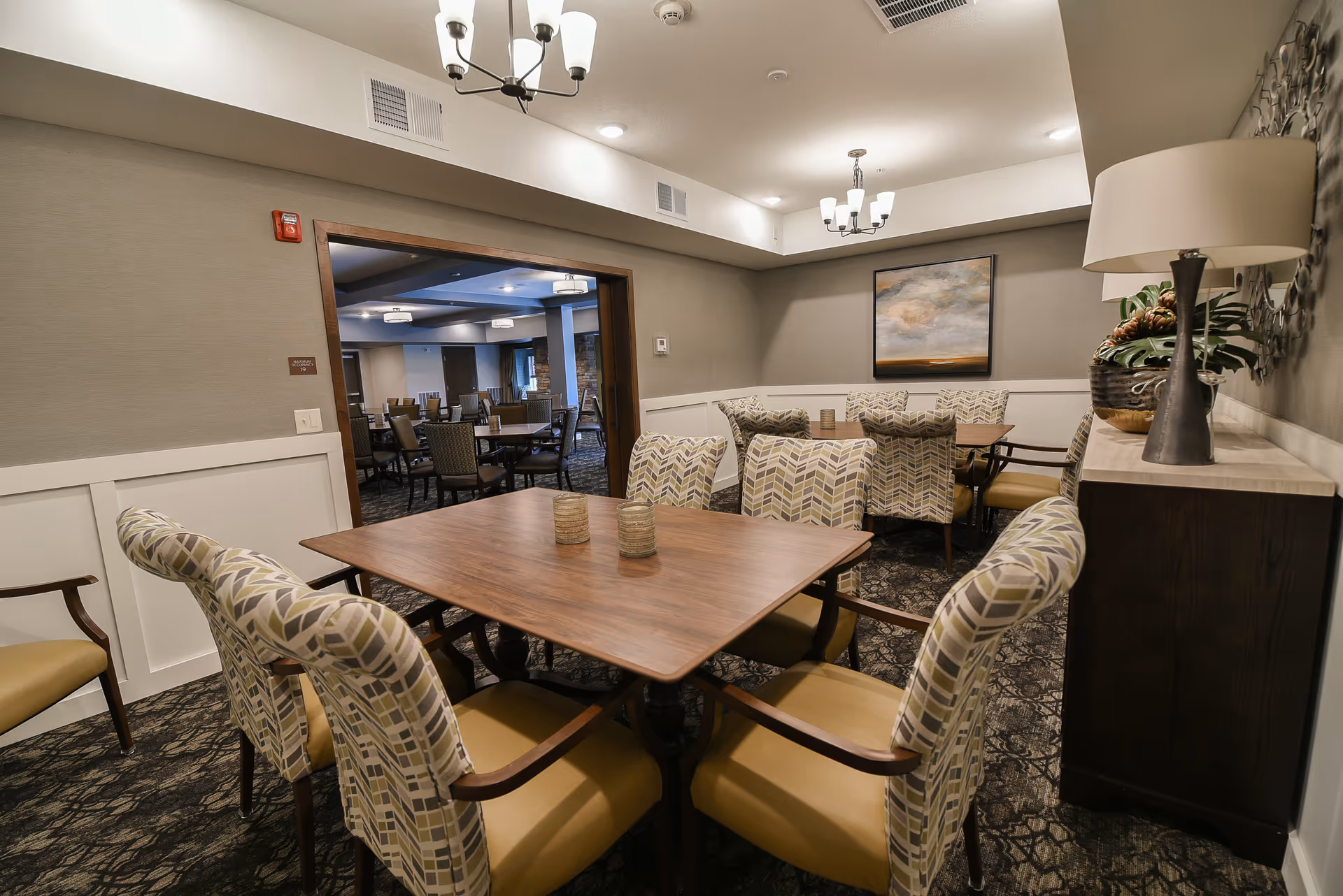 A well-lit communal dining room with wooden tables, patterned upholstered chairs, a sideboard with a lamp, and a painting on the wall.
