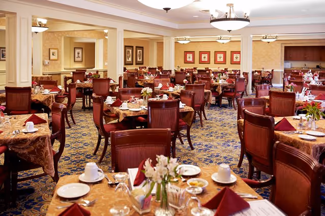 A spacious dining room with multiple tables covered in patterned tablecloths, each set with white plates, cups, silverware, and folded burgundy napkins. The room features wooden chairs, floral centerpieces on the tables, patterned carpet, and warm lighting from ceiling fixtures.