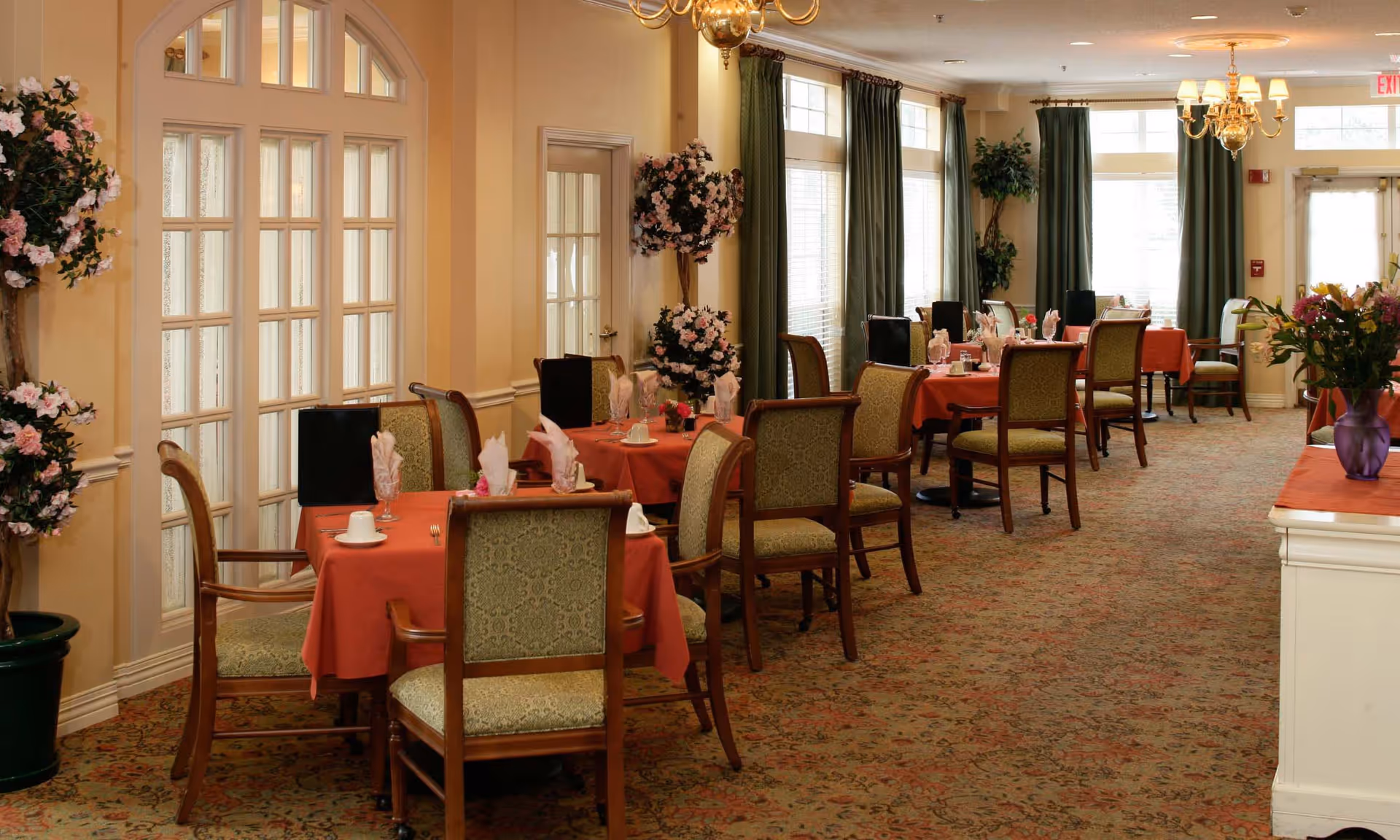 A dining room with several tables covered in red tablecloths, each set with napkins, cups, and utensils. The room features patterned carpet, green curtains on large windows, floral arrangements, and warm lighting from chandeliers.
