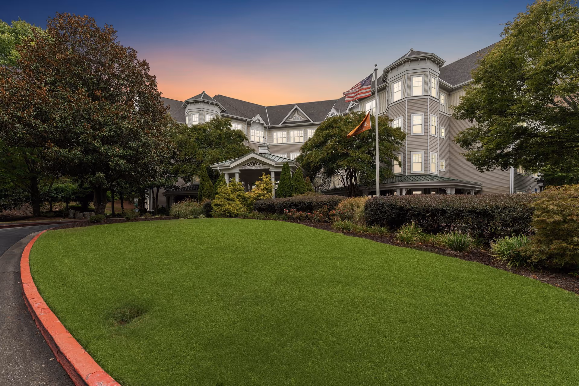 Exterior view of a large senior living facility building at sunset with well-maintained green lawn, trees, shrubs, and two flagpoles displaying the American flag and an orange flag.