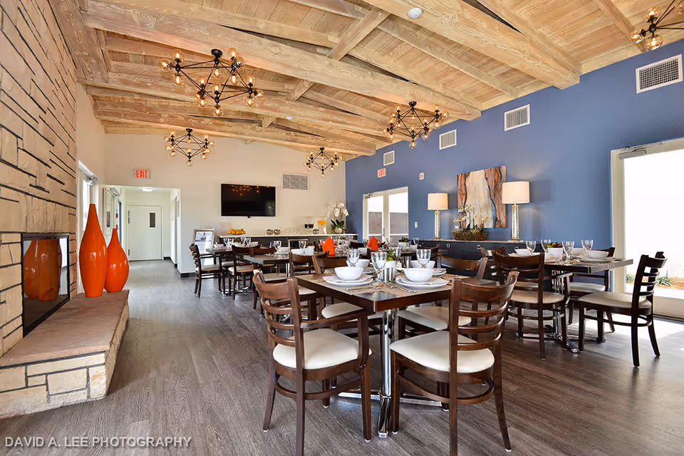 Spacious dining room with wooden tables and chairs, set place settings, a stone fireplace, chandeliers, and a blue accent wall.