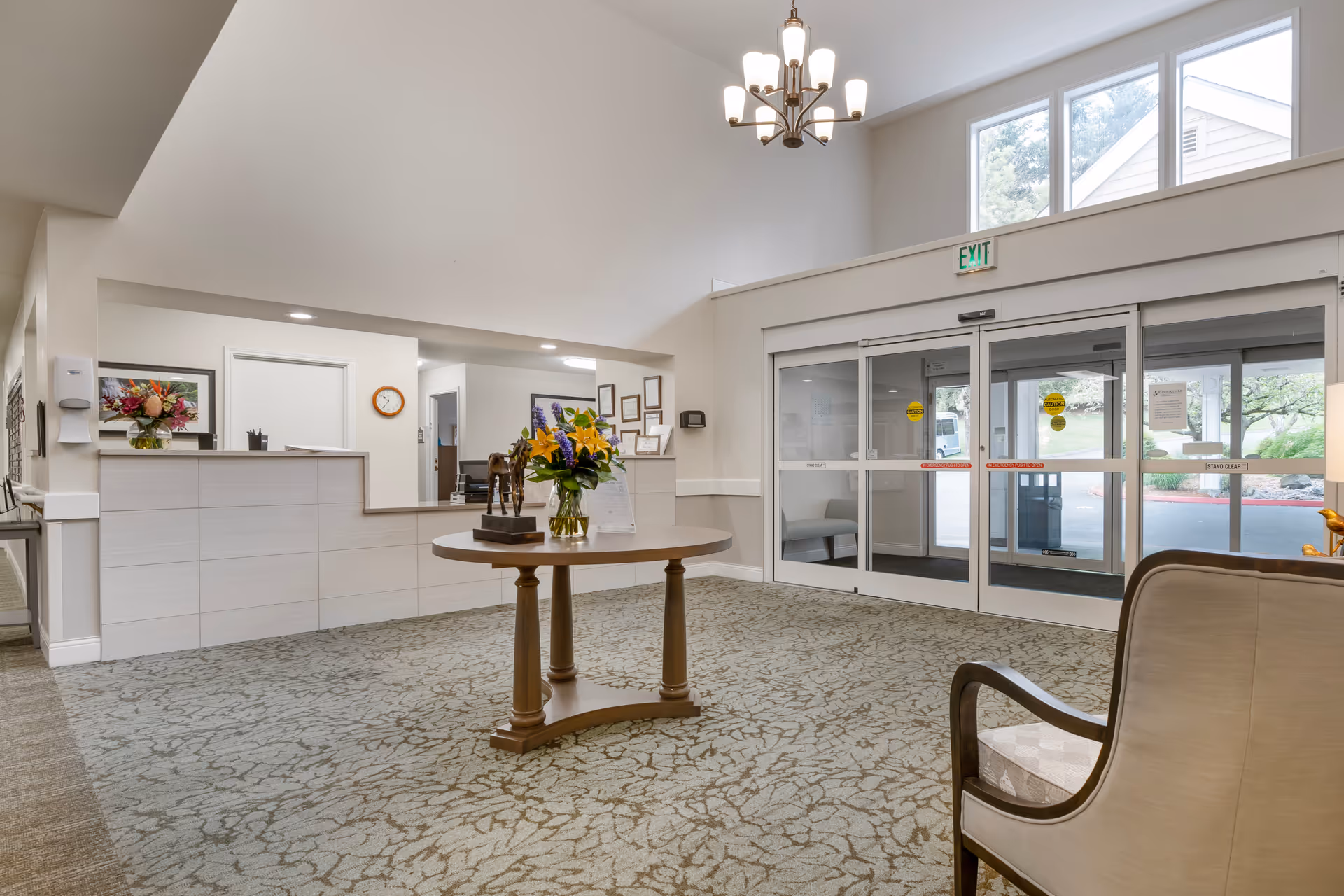 Interior view of a senior living facility lobby with a round wooden table in the center holding a flower vase and a small horse sculpture. To the left is a reception desk with flowers and office supplies. Large glass automatic sliding doors lead outside, with an exit sign above. The space has high ceilings, carpeted floors, and a chandelier light fixture.