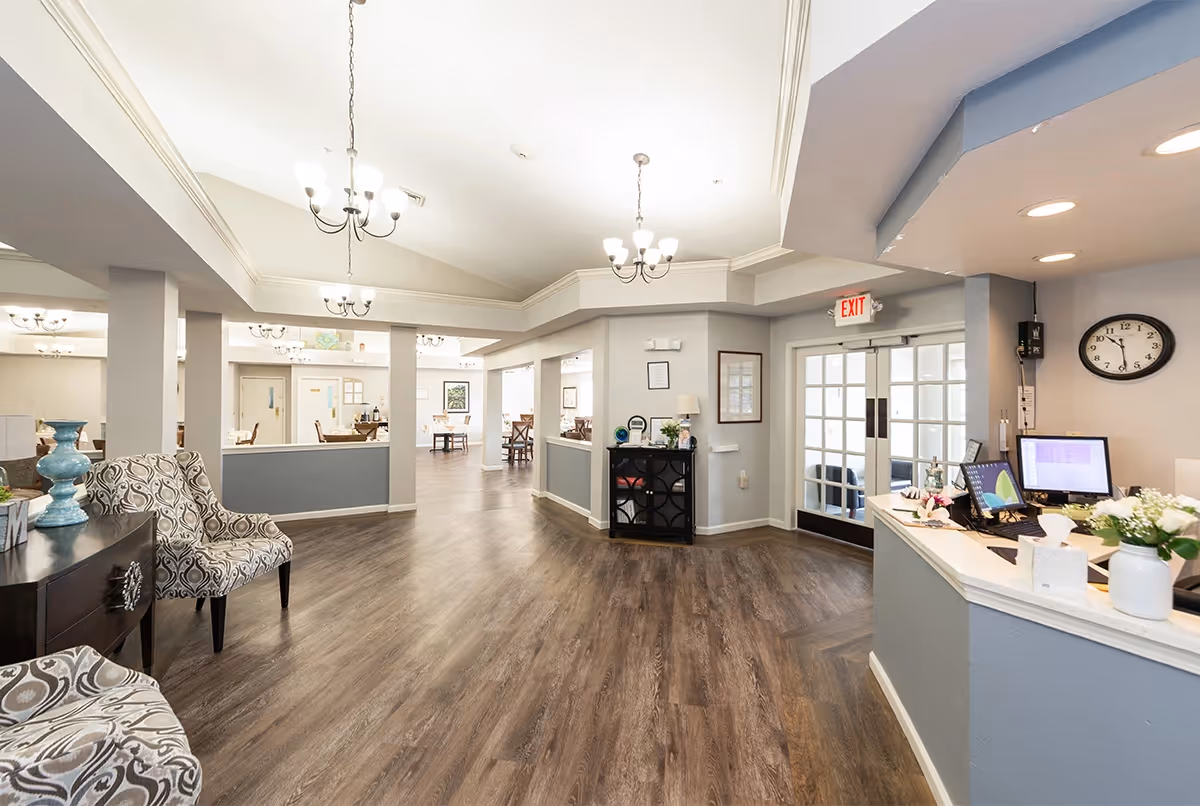 Bright and spacious interior of an assisted living facility with wood flooring, patterned armchairs, a reception desk with computer monitors, and multiple chandeliers hanging from the ceiling. There are glass double doors with an exit sign above them and various tables and chairs visible in the background.
