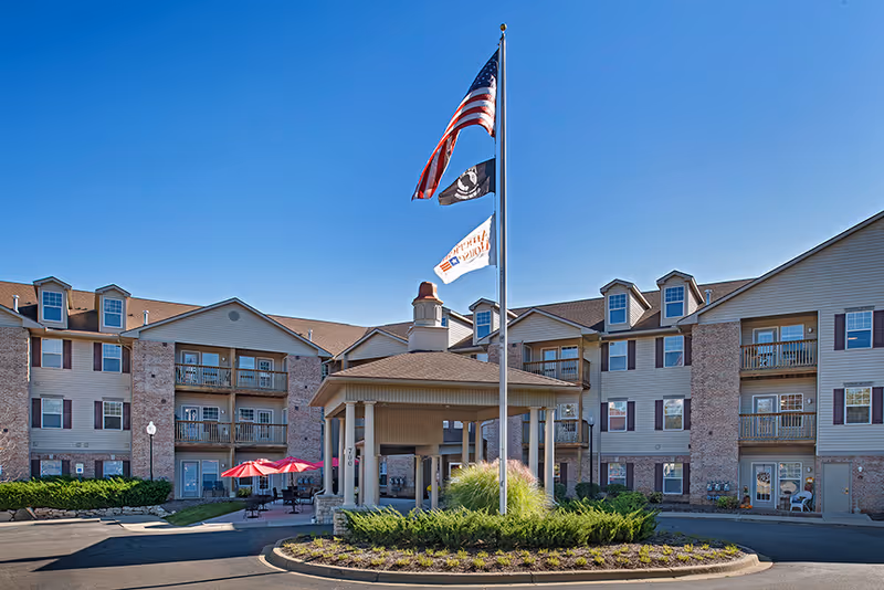 Front exterior of a three-story senior living building with a covered porte-cochere, flagpoles flying flags, balconies, and outdoor seating with red umbrellas.