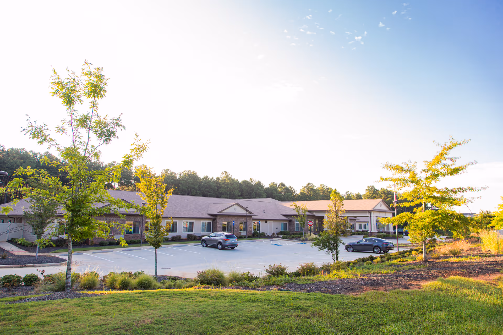 Exterior view of a single-story assisted living and memory care facility building with a parking lot in front, two cars parked, several young trees, and a grassy area in the foreground under a clear sky.