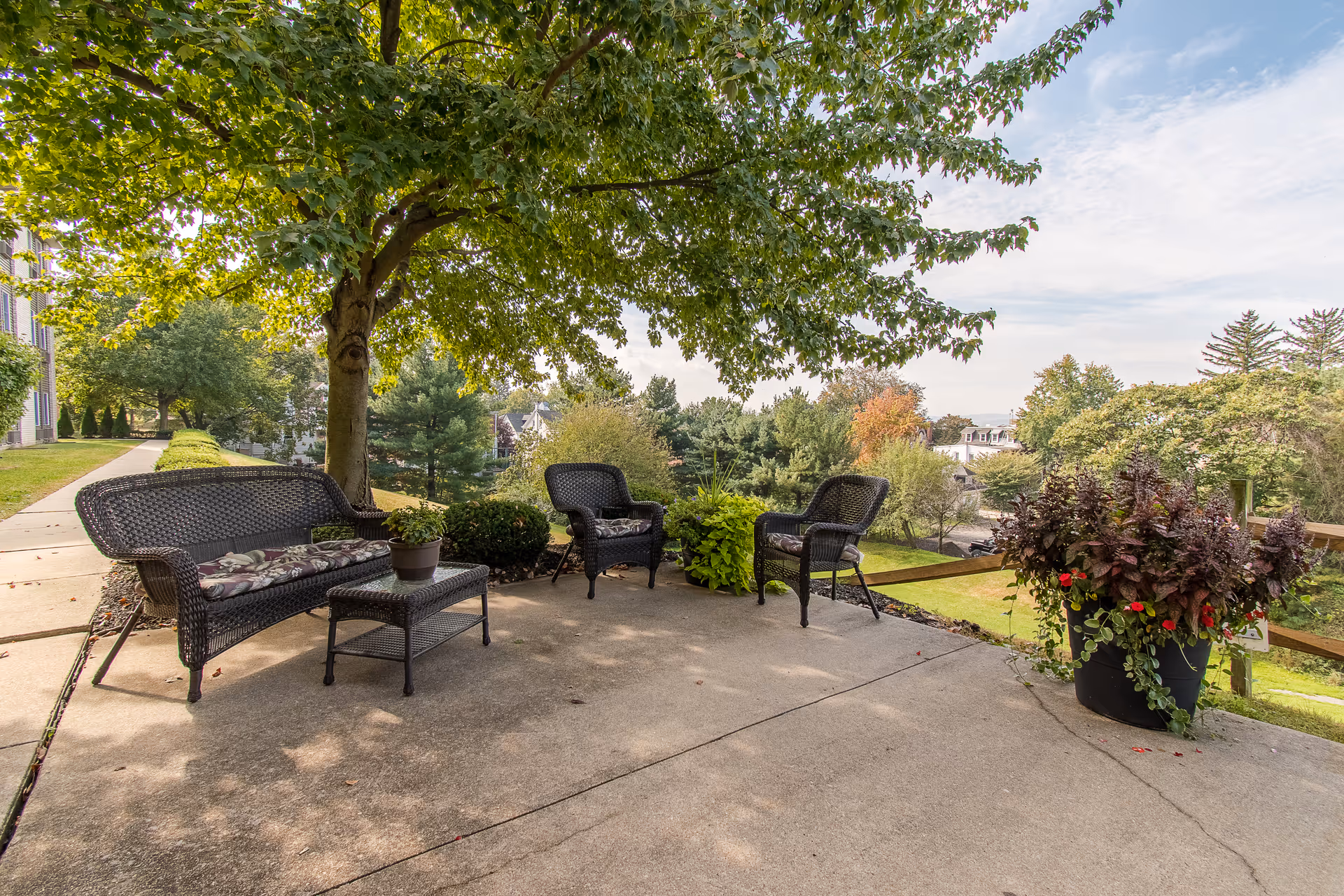 Shaded outdoor patio with wicker bench, chairs and a small table under a large tree overlooking a landscaped lawn.