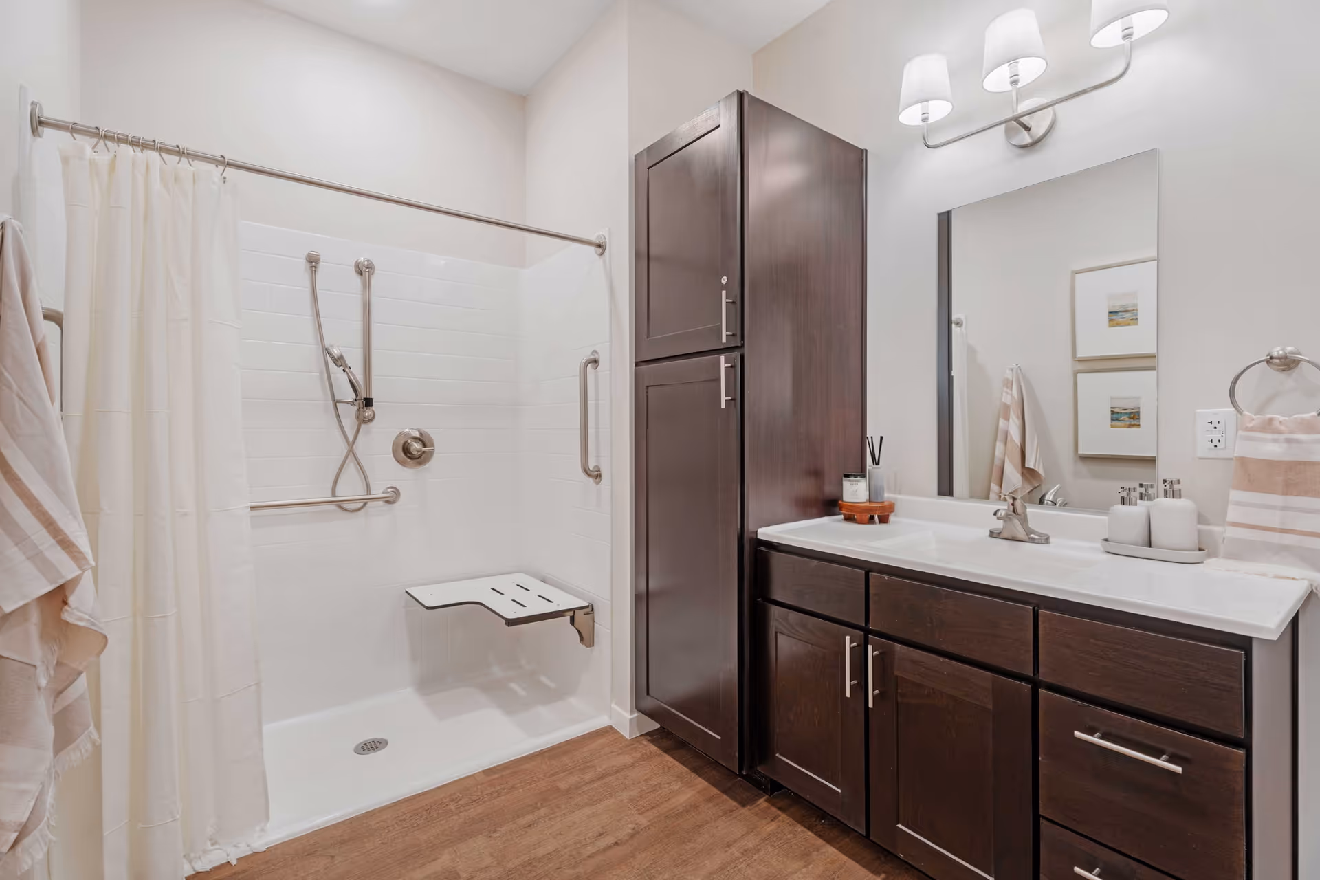 A modern bathroom featuring a walk-in shower with white tiles, a fold-down shower seat, and grab bars. There is a shower curtain on a rod, dark wood cabinetry with a white countertop, a sink with a faucet, a large mirror, and wall-mounted light fixtures above the mirror. Towels hang on a ring and a hook on the walls.