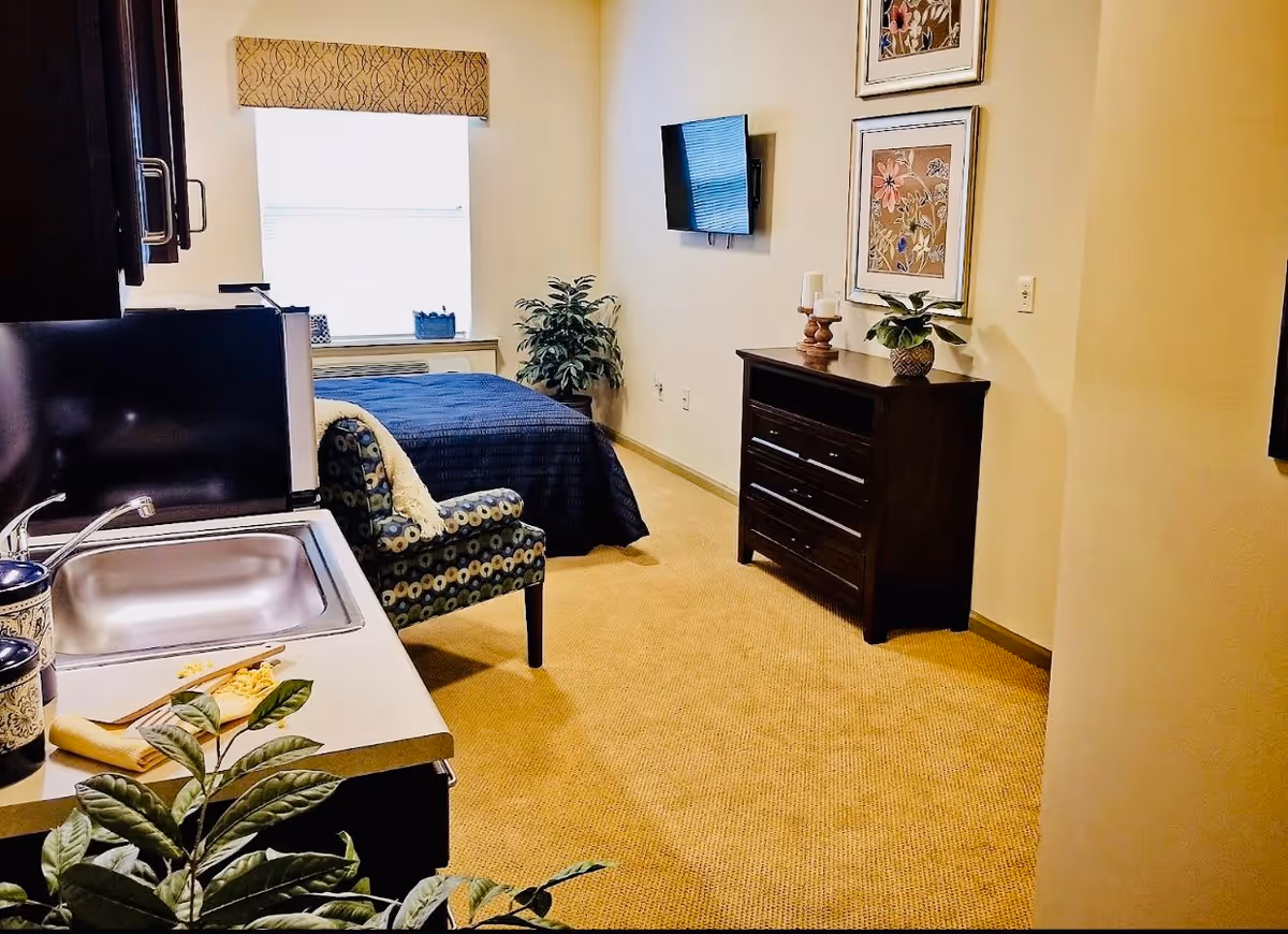 Interior view of a senior living facility room featuring a small kitchenette with a sink and microwave on the left, a patterned armchair, a bed with a dark blue cover near a window with a valance, a dark wooden dresser with decorative items, a wall-mounted TV, and framed floral artwork on the wall.