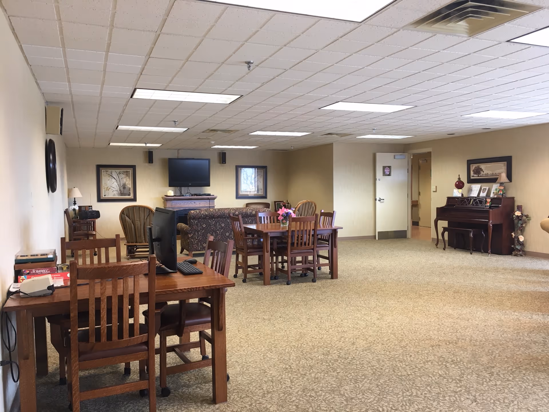 A spacious interior common area with wood tables, chairs, piano, and a wall-mounted TV