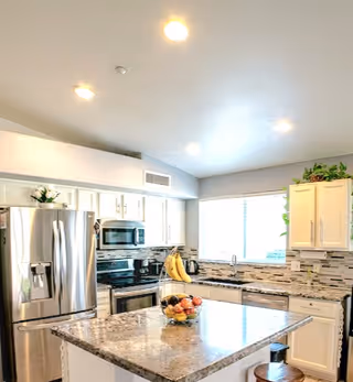 Bright modern kitchen with white cabinets, stainless steel refrigerator, oven, and microwave. A large granite island with a bowl of fruit and bananas on a stand is in the center. There is a window above the sink letting in natural light and some green plants on top of the cabinets.