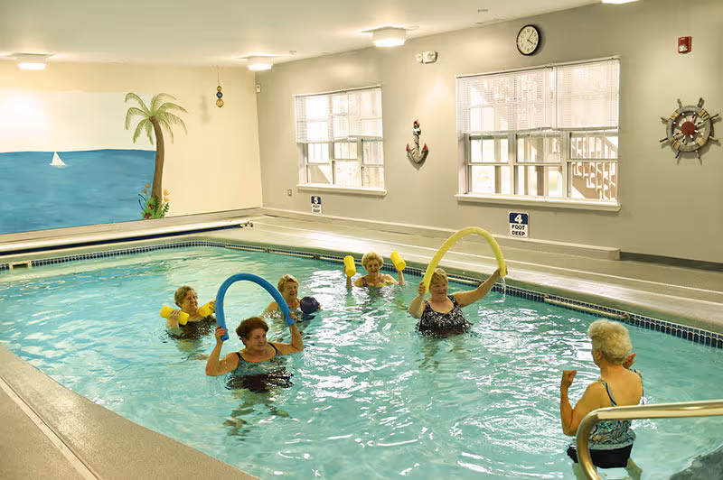 Several senior residents doing water exercise with pool noodles in an indoor swimming pool with nautical decor.