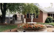 Outdoor courtyard area with a circular brick planter filled with flowers and a white decorative fountain in the center. Surrounding the planter are trees, shrubs, and a brick building with windows in the background.