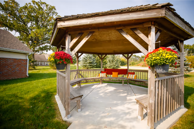 A wooden gazebo with a shingled roof situated on a concrete patio in a grassy outdoor area. The gazebo has built-in benches with colorful cushions and hanging flower pots with red and yellow flowers on the corners. Trees and a brick building are visible in the background under a clear sky.