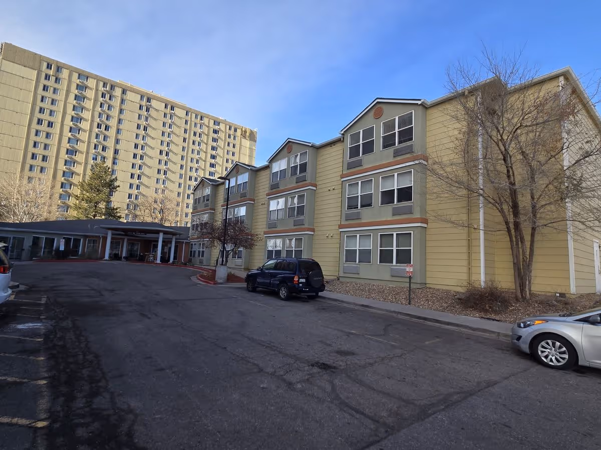 Front exterior of a multi-story senior living building with a covered entrance, driveway, and parked cars.