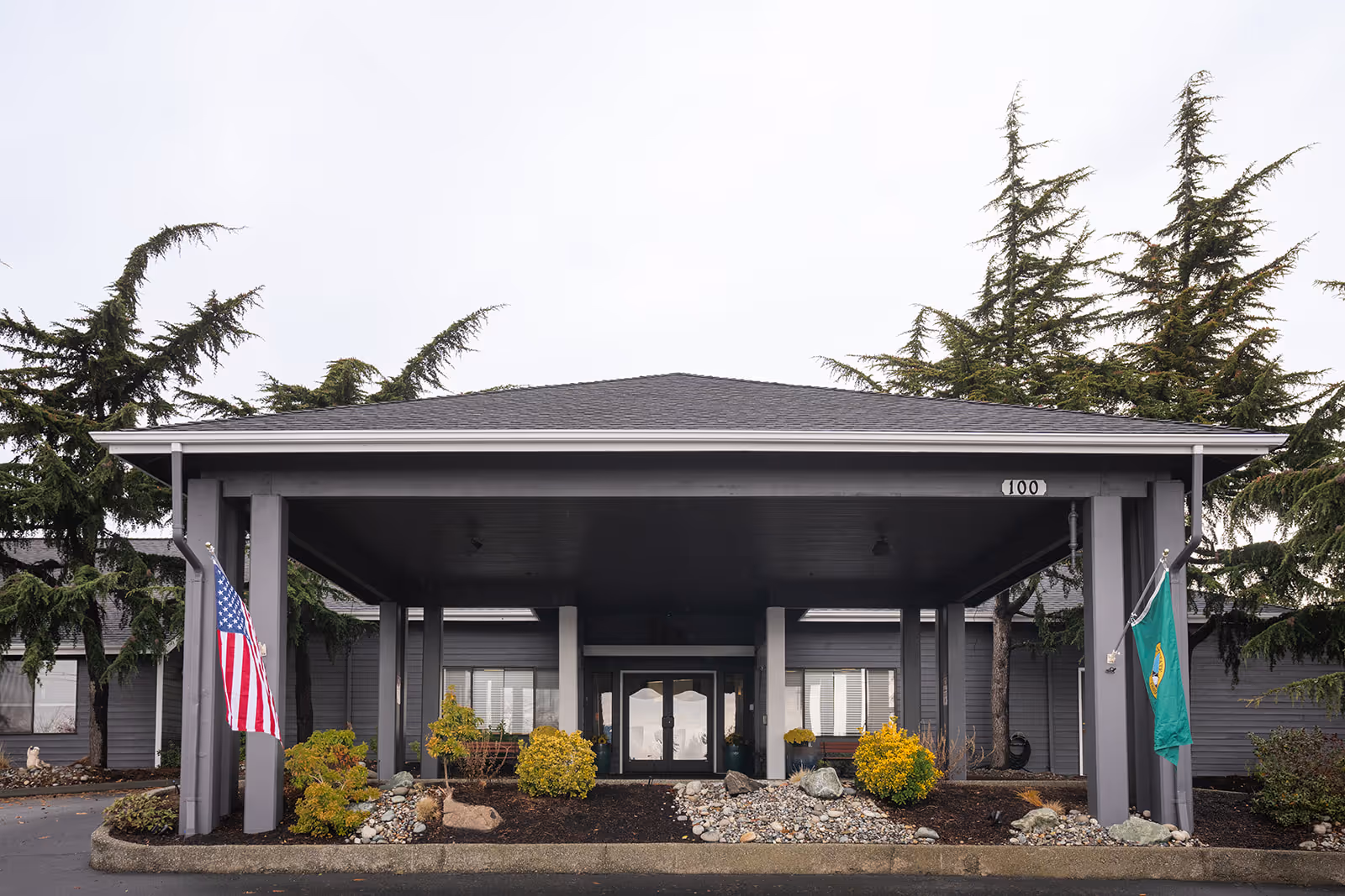 Covered entrance/portico of a gray senior living building with American and Washington state flags and landscaped beds.