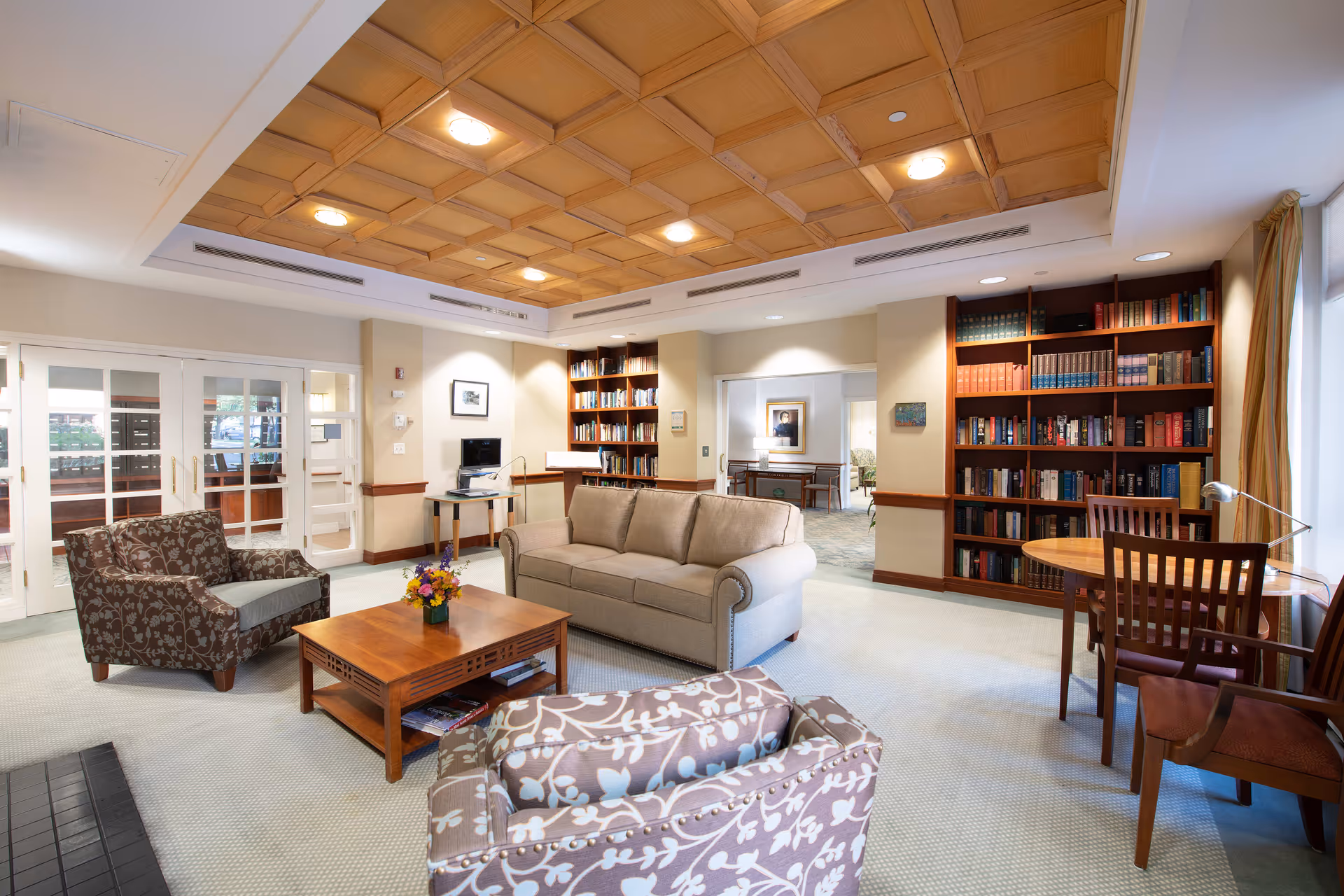 Bright communal living room with sofas, armchairs, bookshelves, a coffee table, and a coffered wooden ceiling.
