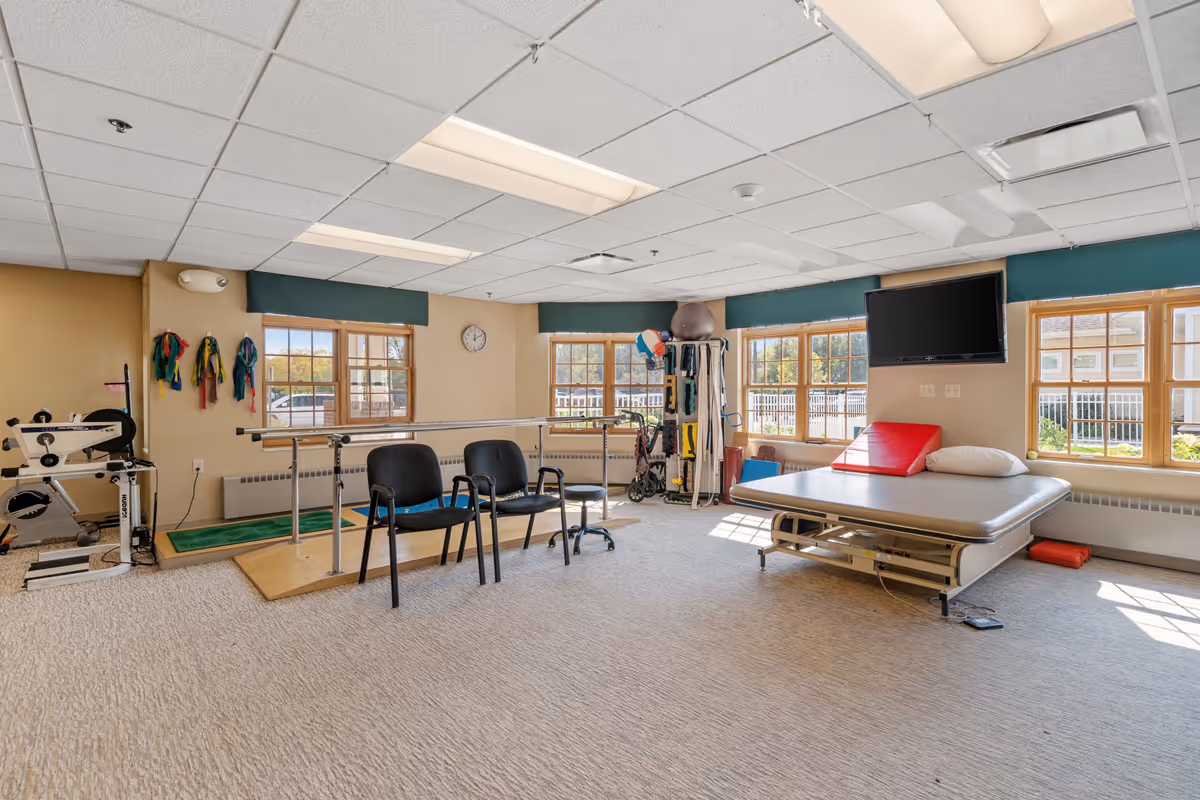 A well-lit therapy or rehabilitation room with exercise equipment including parallel bars, chairs, resistance bands hanging on the wall, a therapy bed with a red cushion and pillow, a TV mounted on the wall, and large windows letting in natural light.