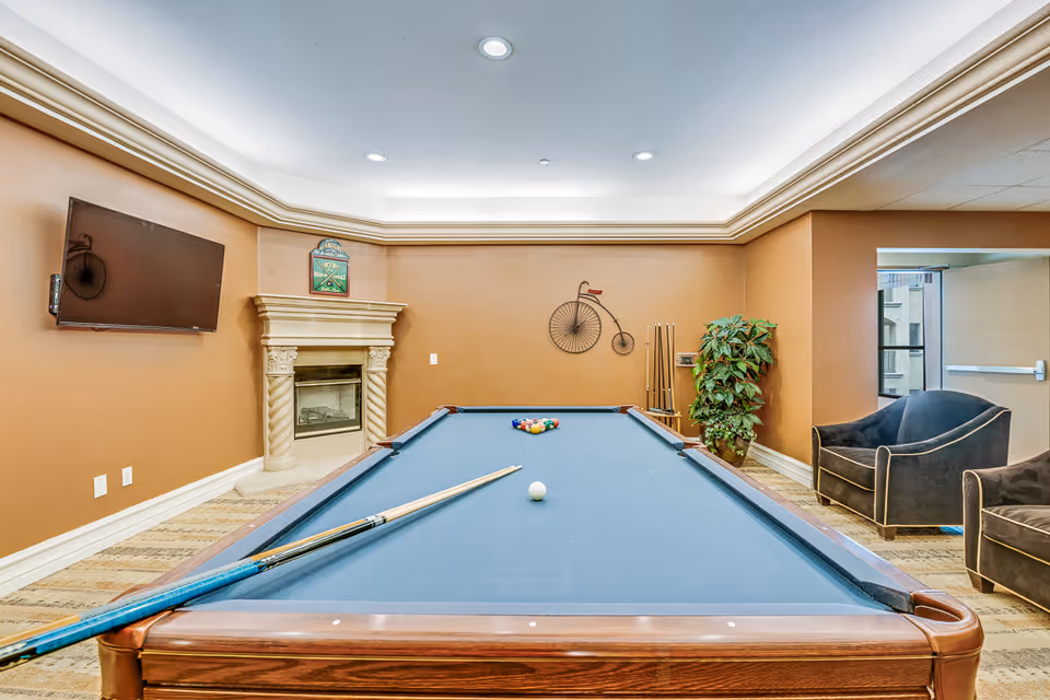 Interior recreation room with a pool table in the foreground, seating, a fireplace, wall-mounted TV, and decorative wall art.