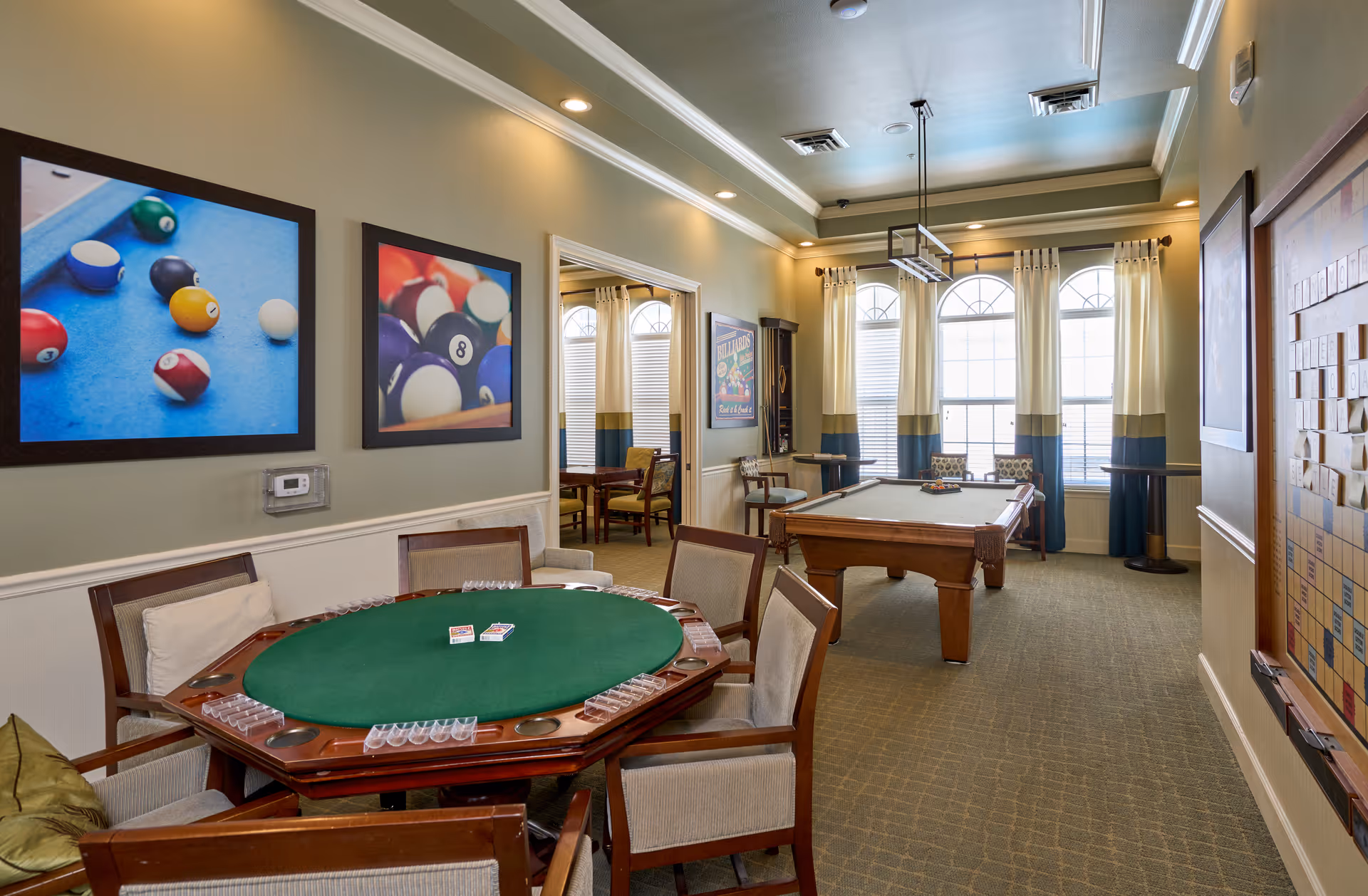 A well-lit game room featuring a green felt poker table with chairs around it, a pool table with billiard balls arranged, and framed pictures of billiard balls on the walls. The room has large windows with curtains, carpeted floor, and a Scrabble board mounted on the wall.