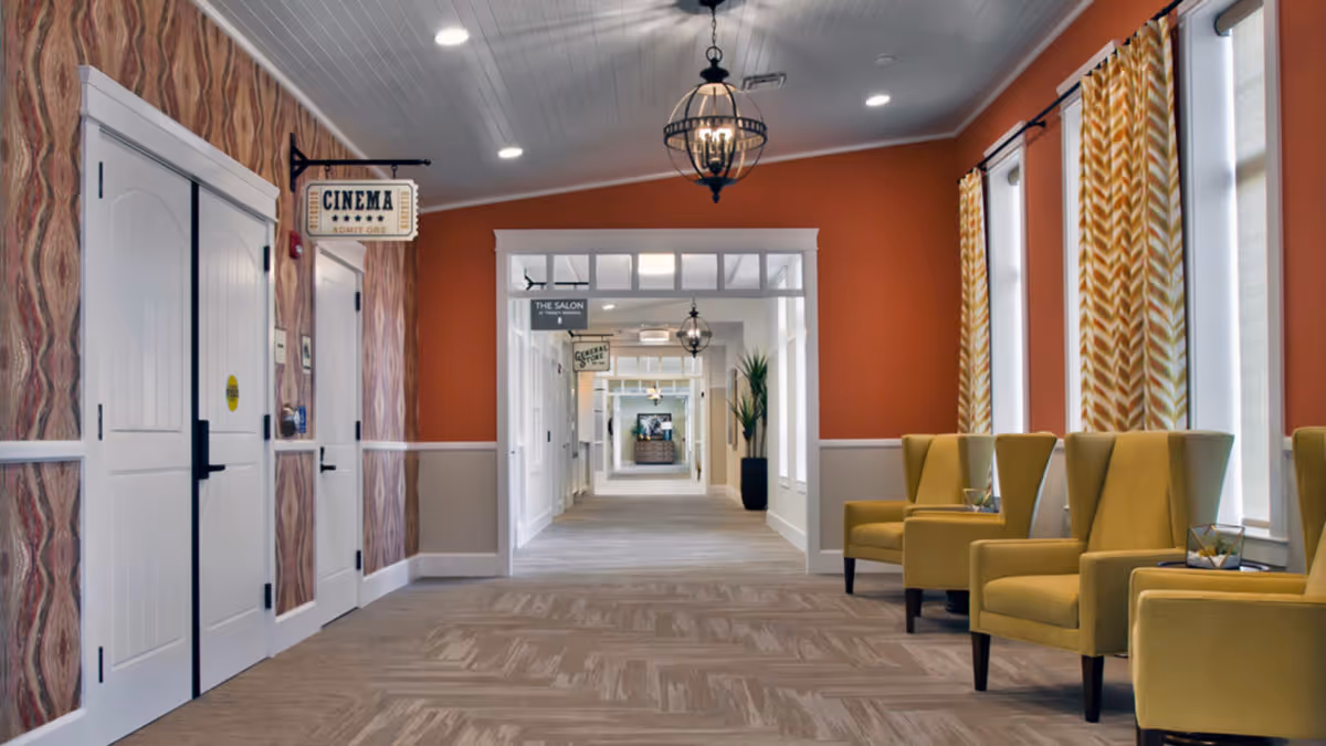 Bright interior corridor with a row of yellow armchairs by windows, decorative wallpaper, ceiling lights, and signs for 'Cinema' and 'The Salon'.