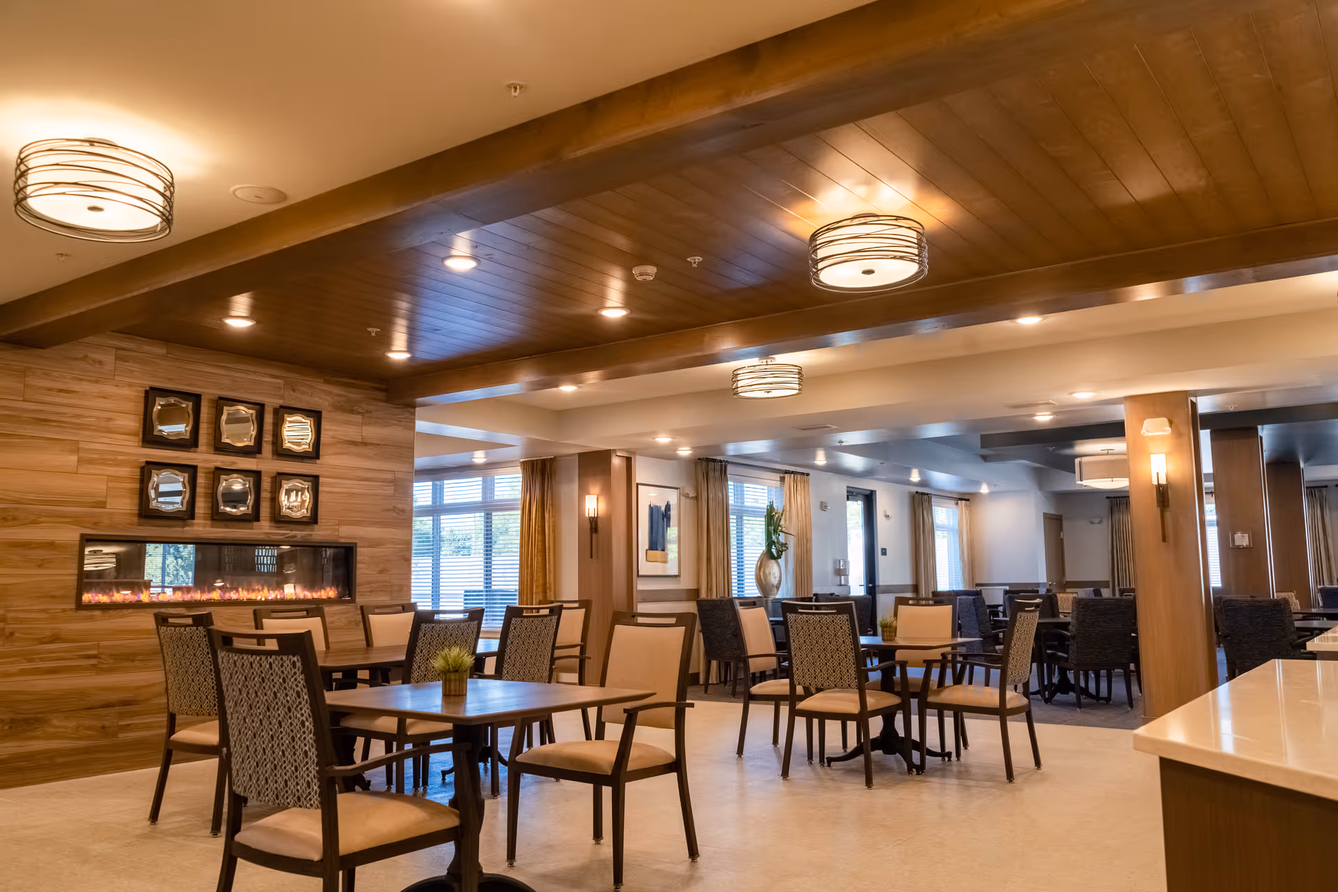 A spacious dining room in a senior living facility with multiple tables and chairs arranged neatly. The room features a modern electric fireplace set into a wooden accent wall, decorative framed mirrors above the fireplace, large windows with curtains allowing natural light, and warm ceiling lights with wooden beams.