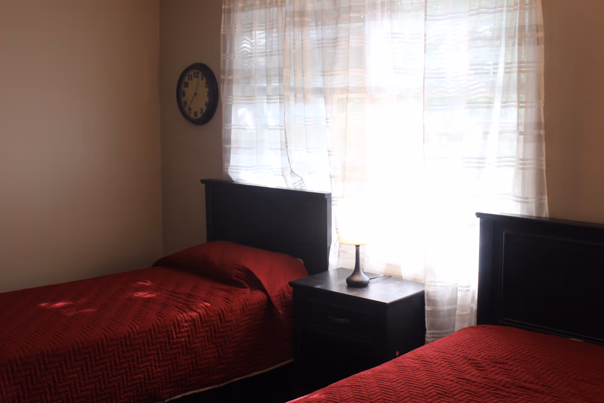 Two twin beds with red bedspreads flanking a nightstand and lamp in a softly lit bedroom with sheer curtains and a wall clock.