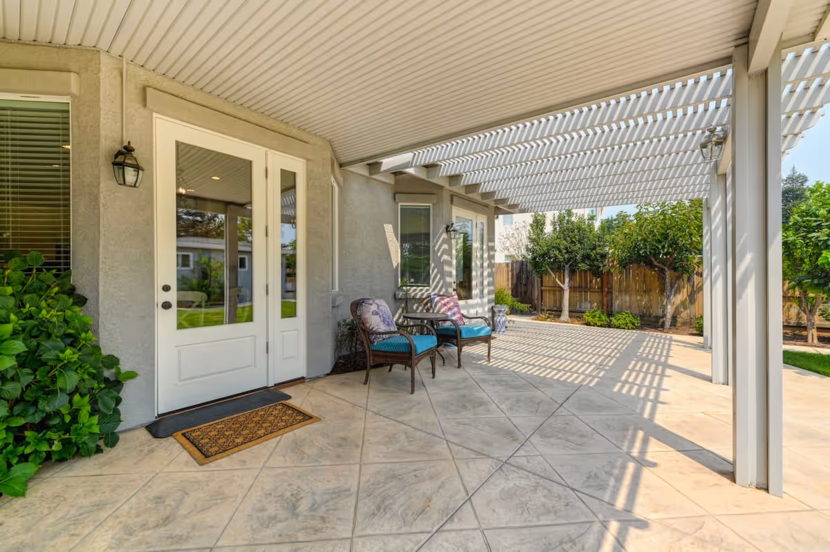 Outdoor patio area with a white pergola casting striped shadows on the tiled floor. Two cushioned chairs with floral pillows and a small table are placed near a set of white double doors leading inside. Green plants and trees are visible along the fenced backyard.