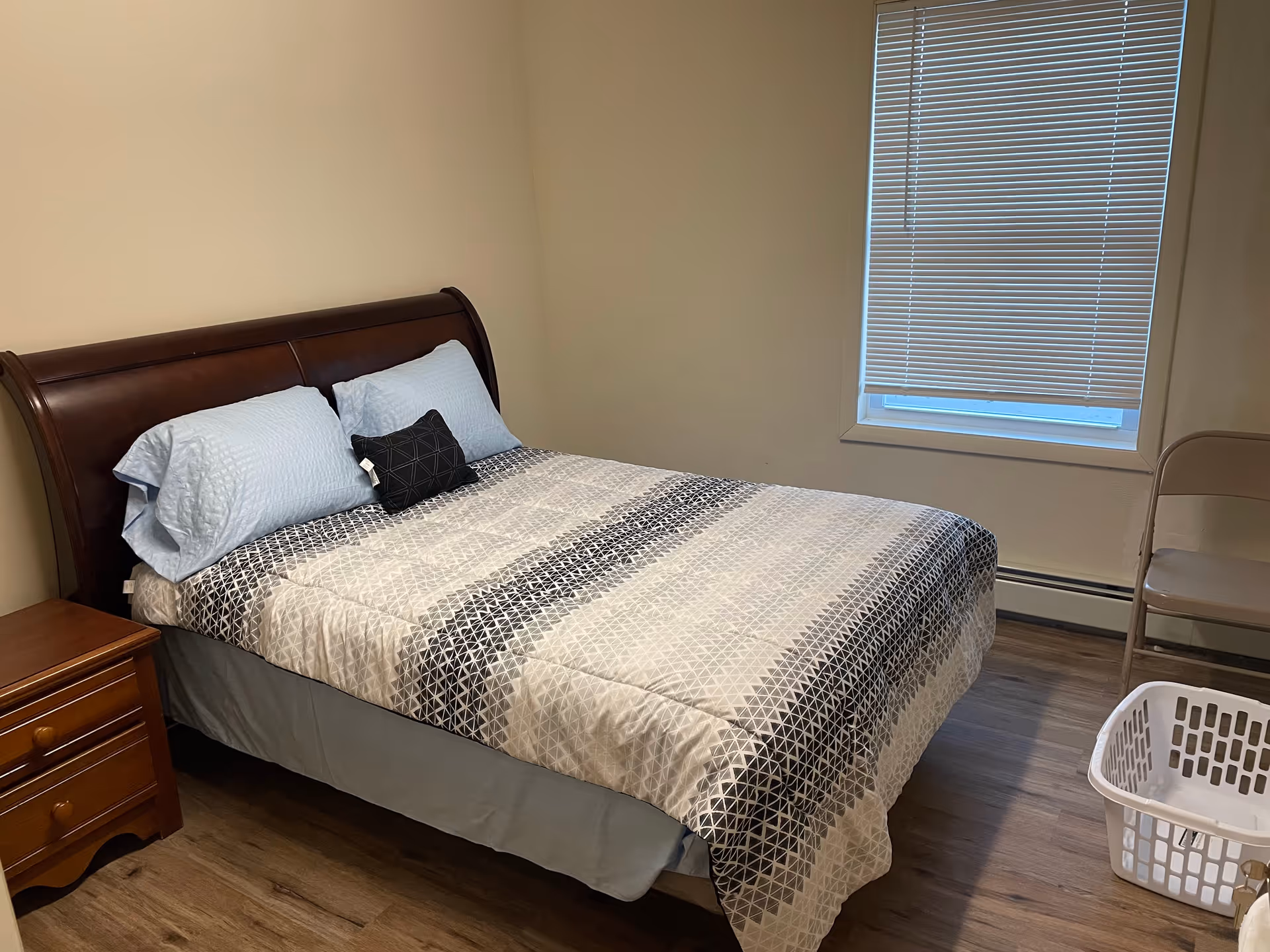 A bedroom with a wooden bed frame and a neatly made bed with patterned bedding and pillows. There is a wooden nightstand with drawers to the left of the bed, a window with closed blinds on the right wall, a folding chair, and a white laundry basket on the floor.