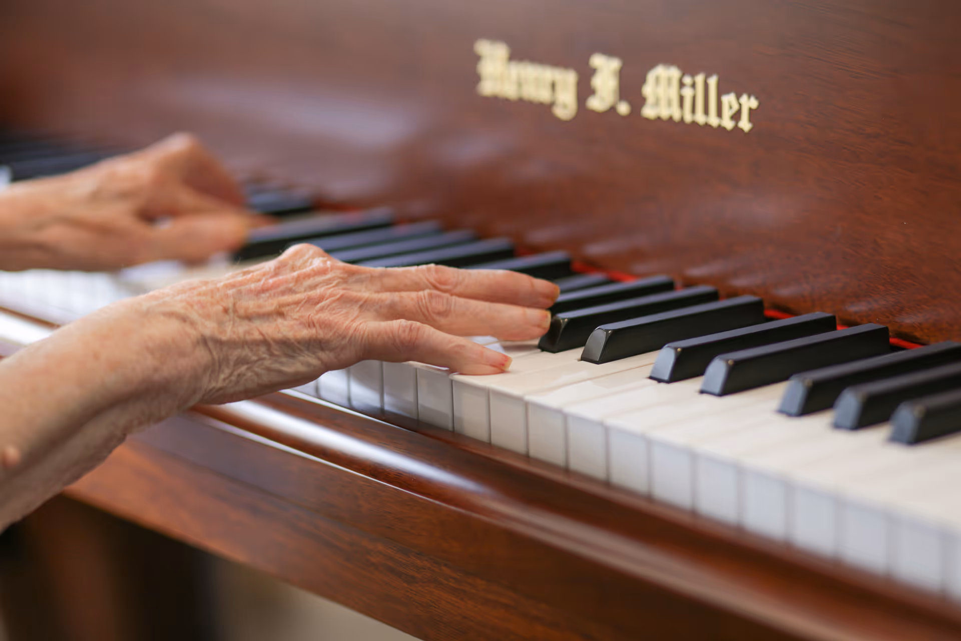 Close-up of elderly hands playing a wooden piano with the name Harry J. Miller engraved above the keys.