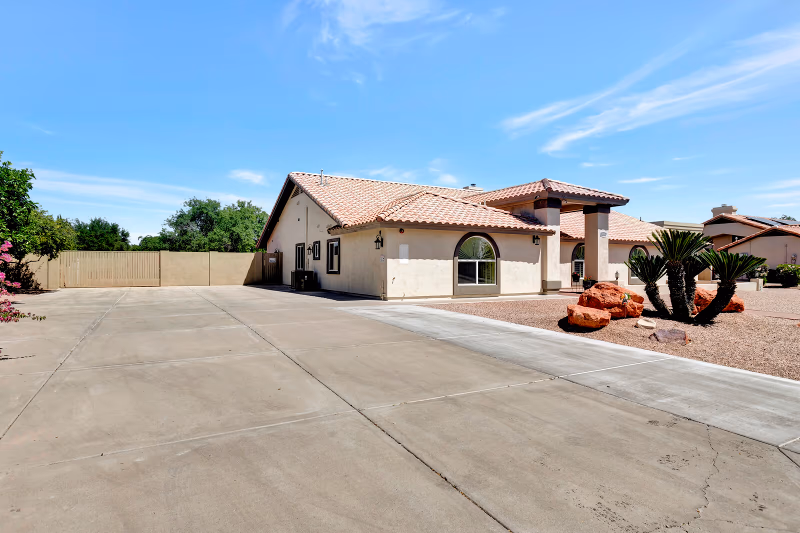 Exterior view of a single-story building with a tiled roof and arched windows, surrounded by a concrete driveway and desert landscaping with rocks and small palm plants under a clear blue sky.