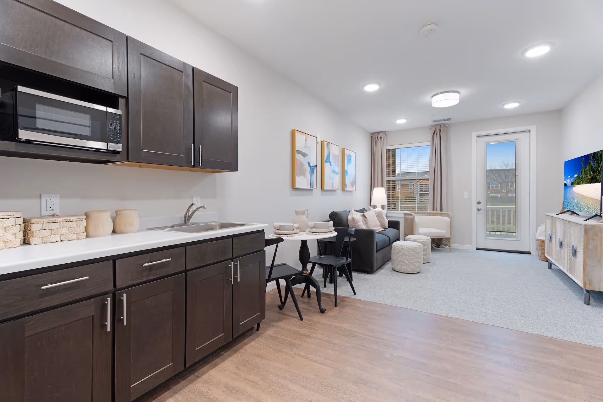 A modern senior living apartment interior featuring a kitchenette with dark wood cabinets, a microwave, and a sink on the left. Adjacent to the kitchenette is a small round dining table set for two with black chairs. The living area includes a dark sofa with light-colored pillows, two small round ottomans, a light armchair, a floor lamp, and three abstract paintings on the wall. A large window with curtains and a glass door leading outside are visible at the far end. A TV is placed on a wooden console on the right side of the room.