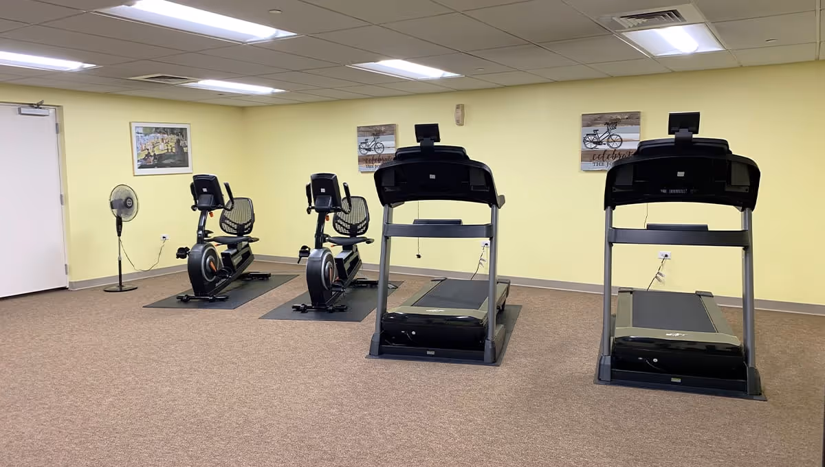 A fitness room with two treadmills and two stationary recumbent bikes placed on mats. The walls are painted light yellow and decorated with framed pictures. There is a standing fan near a closed white door on the left side.