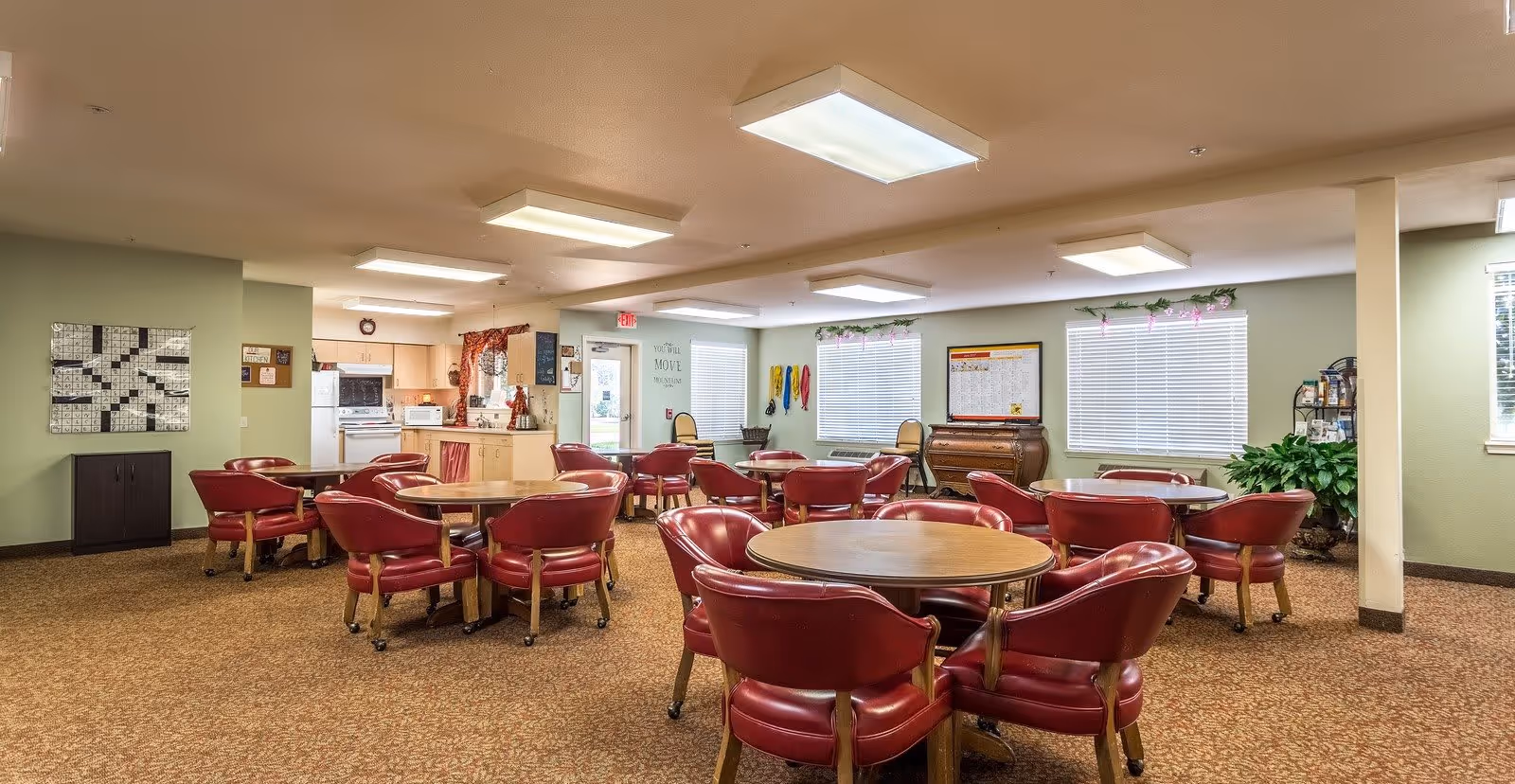 A spacious common dining area with multiple round wooden tables surrounded by red cushioned chairs on wheels. The room has beige carpet flooring, light green walls, and several large windows with white blinds. There is a small kitchen area in the background with white appliances and light wood cabinets. The room is well-lit with rectangular ceiling lights, and there are decorative plants and wall hangings around the space.