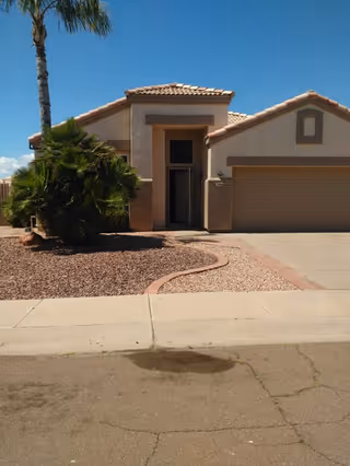 Front exterior view of a single-story house with a tiled roof, a small palm tree and bushes in the front yard covered with gravel, a driveway leading to a two-car garage, and a clear blue sky in the background.
