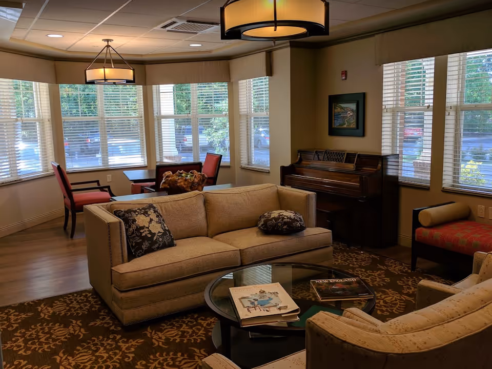 A cozy living room area with a beige sofa and armchair, a glass coffee table with books on it, a wooden piano against the wall, and a small dining table with red cushioned chairs near large windows with white blinds letting in natural light.