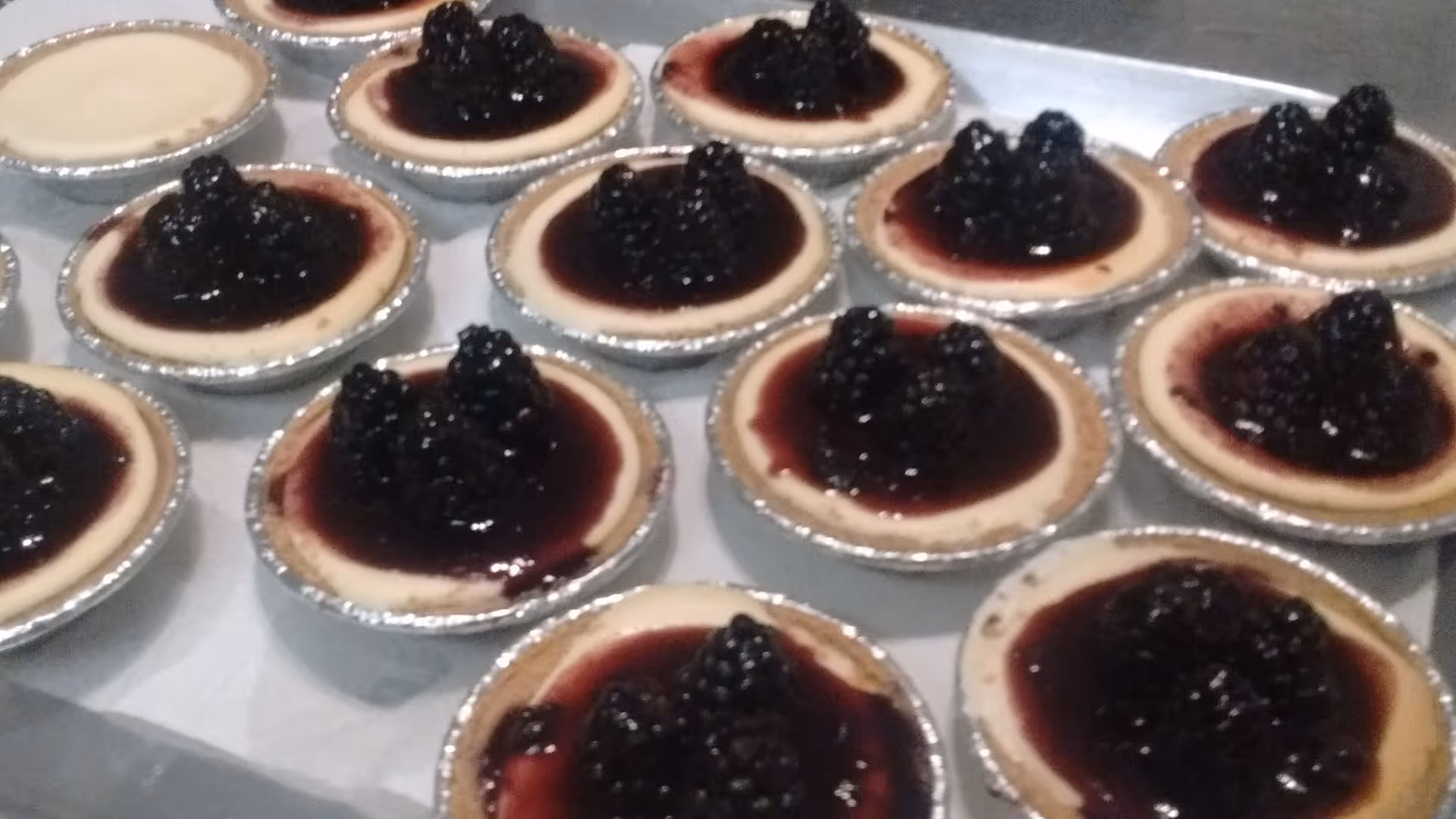 Mini cheesecakes in foil cups topped with blackberry compote on a tray.