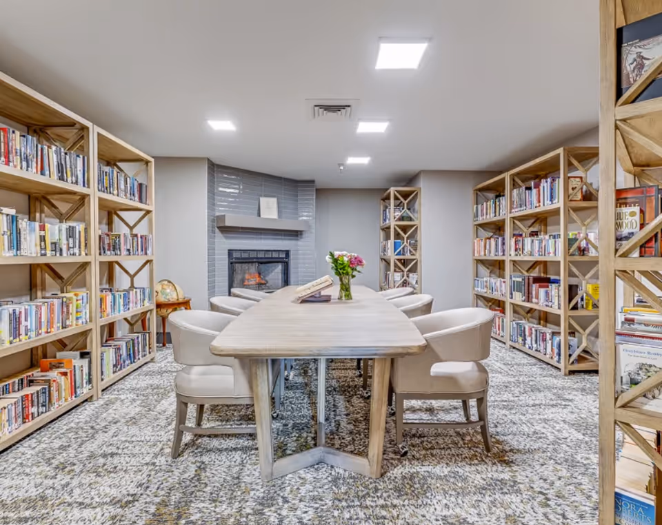 Communal library/reading room with a long wooden table surrounded by chairs, bookshelves along the walls and a fireplace at the far end.