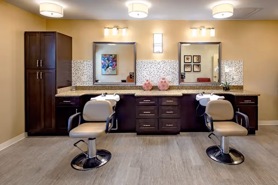 A well-lit salon area with two beige salon chairs in front of a long countertop with two sinks. The countertop has dark wood cabinets and drawers underneath, with mosaic tile backsplash and two large mirrors above. The walls are painted a warm beige color, and there are decorative vases and a small plant on the countertop. The floor has light wood-like flooring.