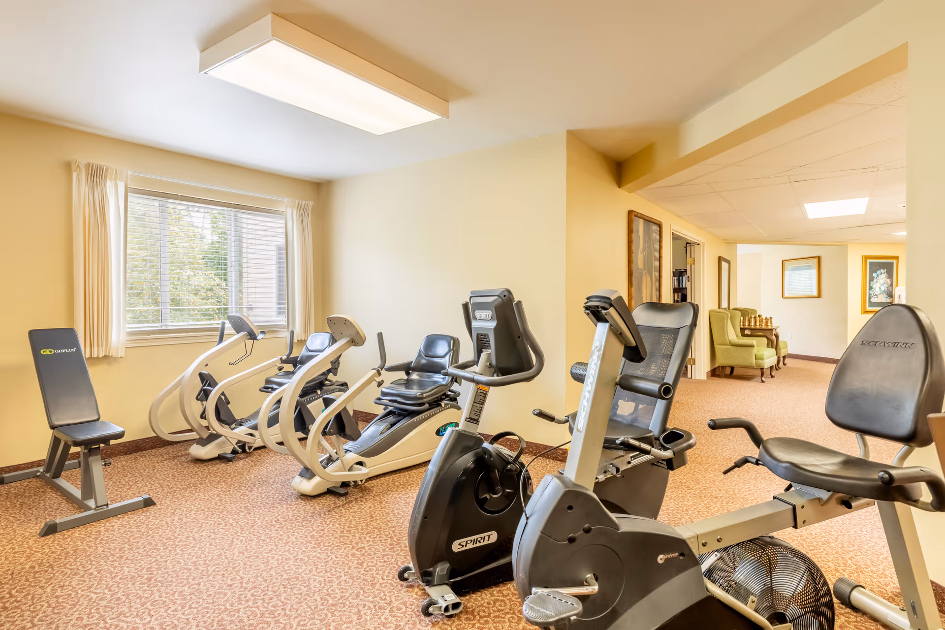 A small fitness room with exercise equipment including stationary recumbent bikes and a weight bench. The room has beige walls, a patterned carpet, a large window with white curtains, and fluorescent ceiling lighting. In the background, there is a hallway with green armchairs and framed artwork on the walls.